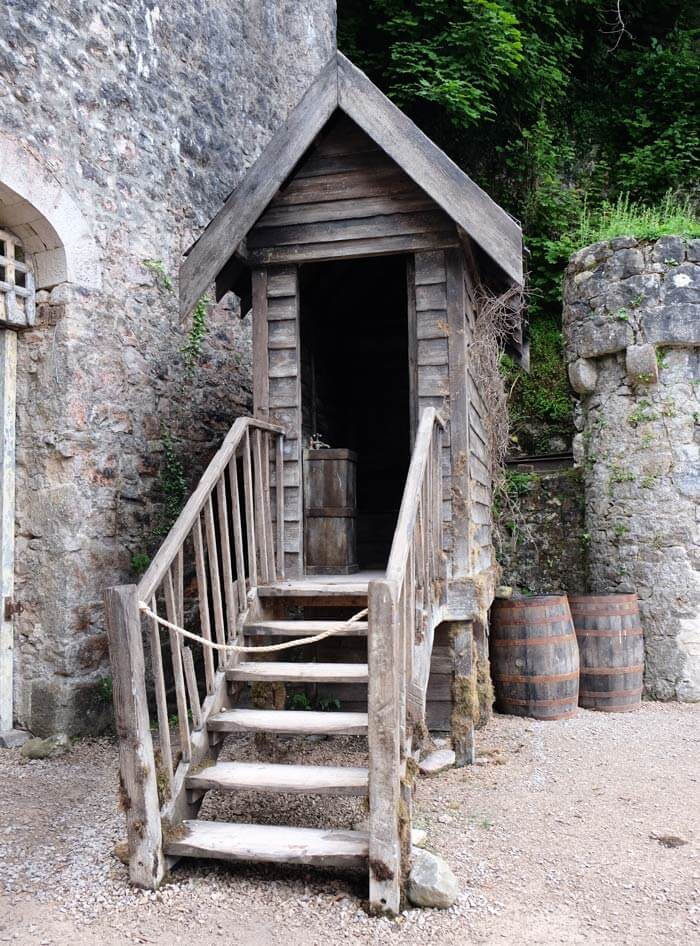 A narrow wooden shed-like building is up some wooden stairs in a castle courtyard. There are stone walls, a small stone tower and some barrels that look like props in the background. 