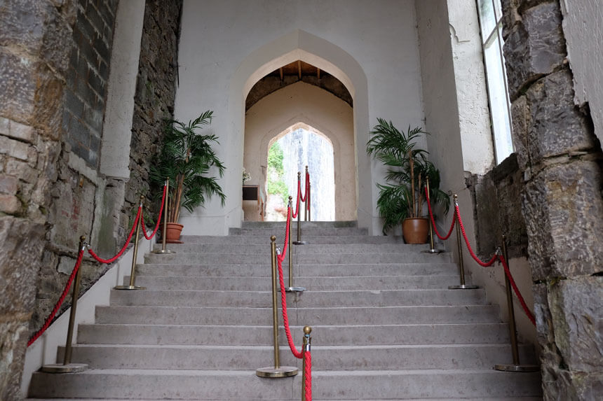 Stone stairs up to the porch at Gwrych Castle. A wide, grand stone staircase with a high, arched ceiling. The walls are partly raw stone, partly plastered. There are two large palm plants and red rope barriers. 