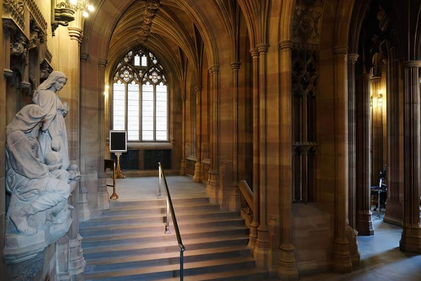 The main entrance hall at the John Rylands library has tall stone columns and a wide stone staircase. It has the air of a cathedral or Hogwarts