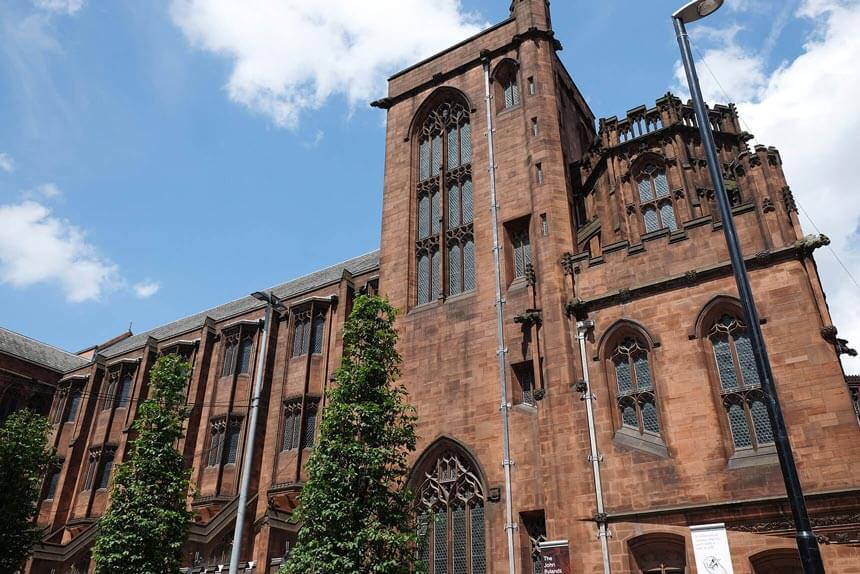 An exterior photo of the John Rylands Library in Manchester. The library is a Gothic style building with arched windows, made of pink stone.