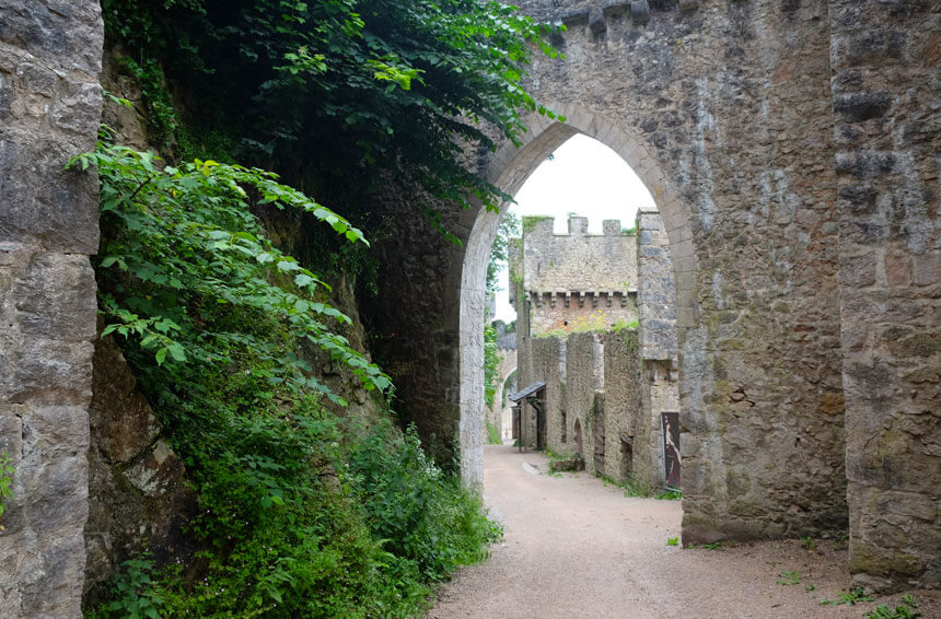 Looking towards the main gate at Gwrych Castle. A narrow lane leads downhill past old stone buildings, a square tower and a circular tower. The walls are rather overgrown and the buildings are in disrepair.