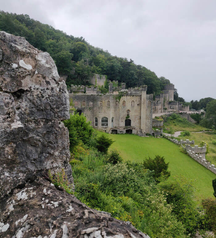 The scene from Nant-y-Bella Tower at Gwrych Castle. A once-grand but ruined Gothic style house with a large grassy lawn in front and woods behind.