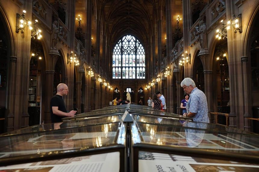 The majestic reading room at the John Rylands Library in Manchester. A large room, with a huge and elaborate stained glass window at one end and lined with stone columns and dim lights. People are looking at displays in glass cases.