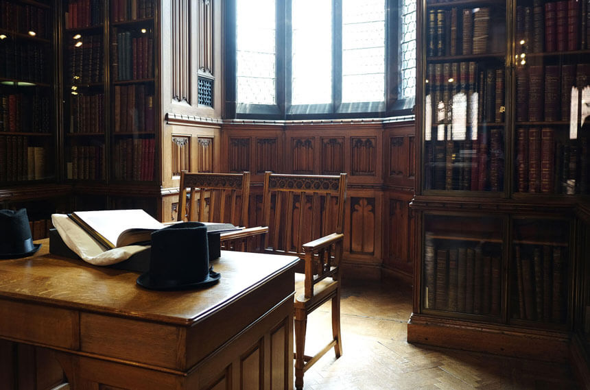 The edges of the John Rylands Library's reading room are lined with wood-panelled study nooks. The nook in the photo has a wooden desk and two wooden chairs. There are bookshelves containing large leather-bound books on either side of the window. 