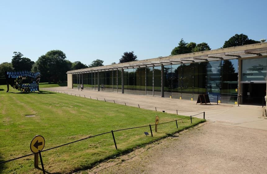 The Underground Gallery at Yorkshire Sculpture Park. A long, glass-fronted modern building with a walkway in front. There is a large sculpture of a mirrored masquerade mask on the grass on the other side of the walkway.