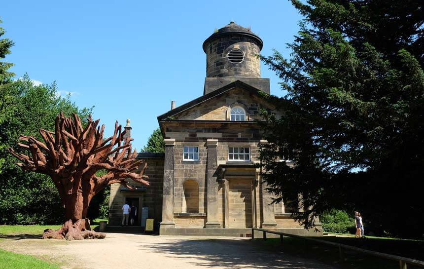 The old Bretton Hall chapel with an iron tree sculpture next to it. The chapel is an old stone building in the classical style, with a circular tower topped with a small dome. There are people entering the chapel gallery on the left hand side and some leaving on the right, walking into the graveyard which is grassy and full of trees.