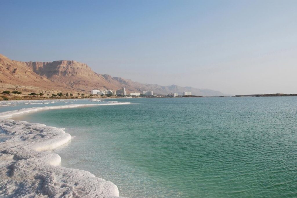 The Dead Sea. A light greeny-blue ocean is edged with salt formations, with sandy-coloured mountains in the background.