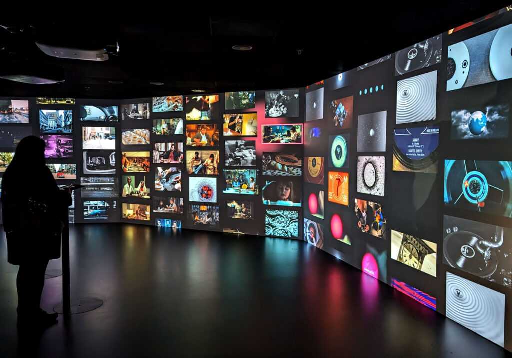 A wall displaying clips of lots of films and TV programmes. A woman is stood on the left, choosing which clip to find out more about next.