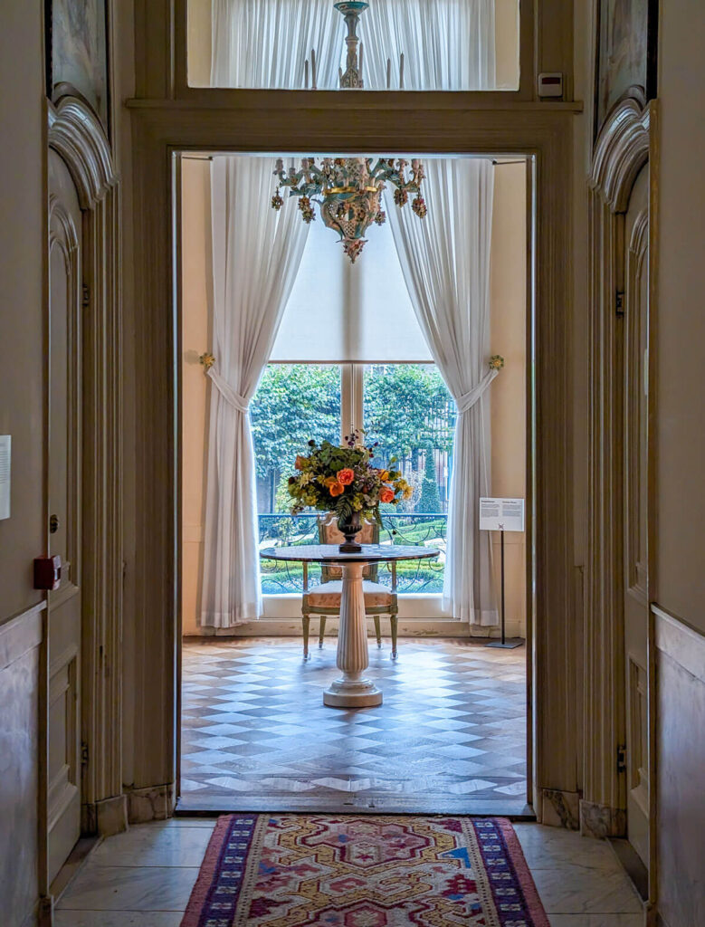 The view through the hallways of the Willet-Holthuysen House. A beautiful hallway in a grand house leads to a table with flowers on it and a window with a view of the garden.