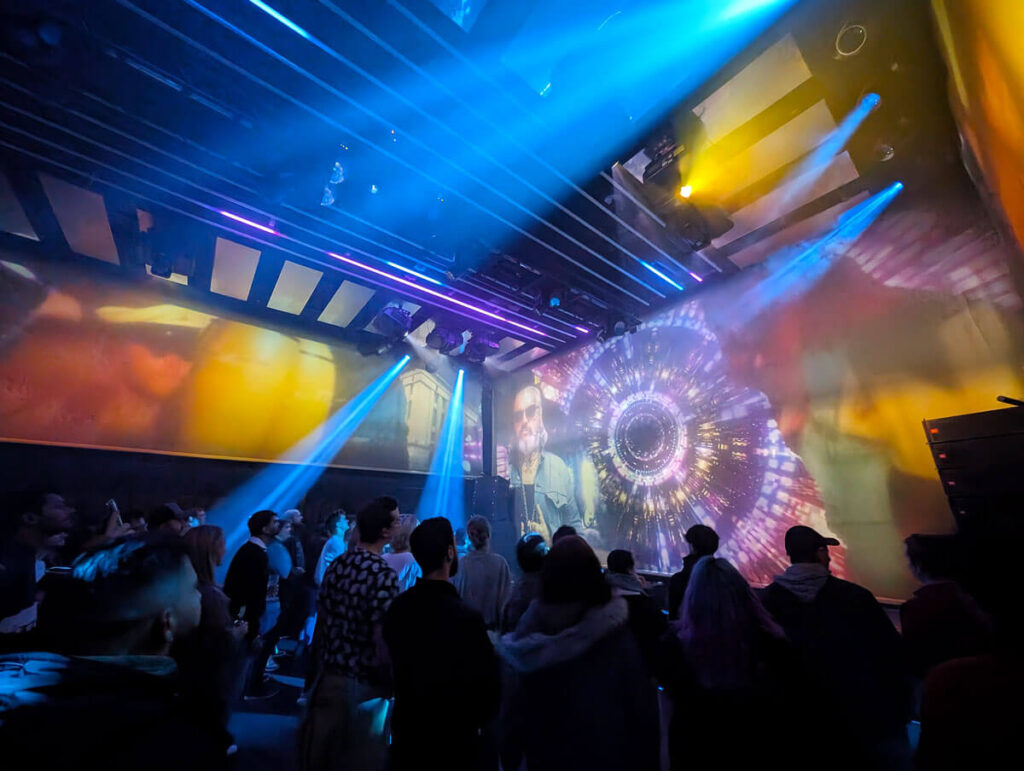 Hitting the dance floor at the end of the Our House experience. A crowd of people are on a dance floor with large screens on two sides and laser lights.