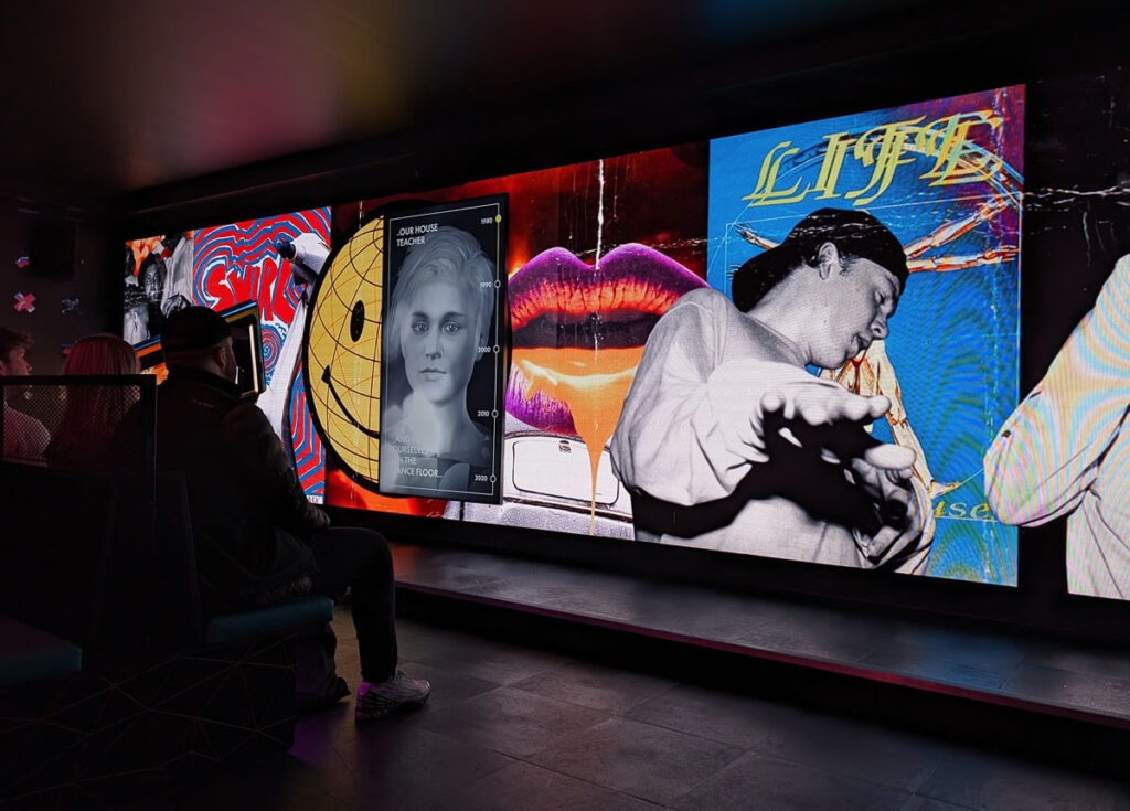 One of the exhibits at Our House. An animated teacher is narrating a video about the history of dance music as people sit down and watch.