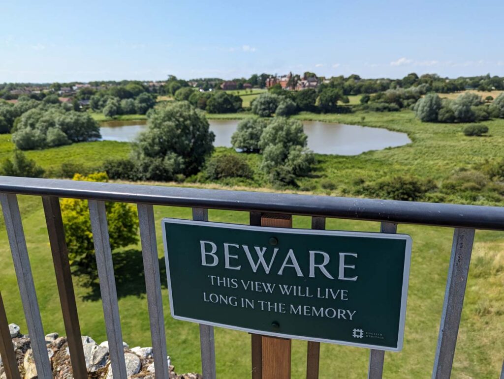 A view from Framlingham Castle's walls. A sign reads "Beware, this view will live long in the memory". The view is of green fields and a lake.