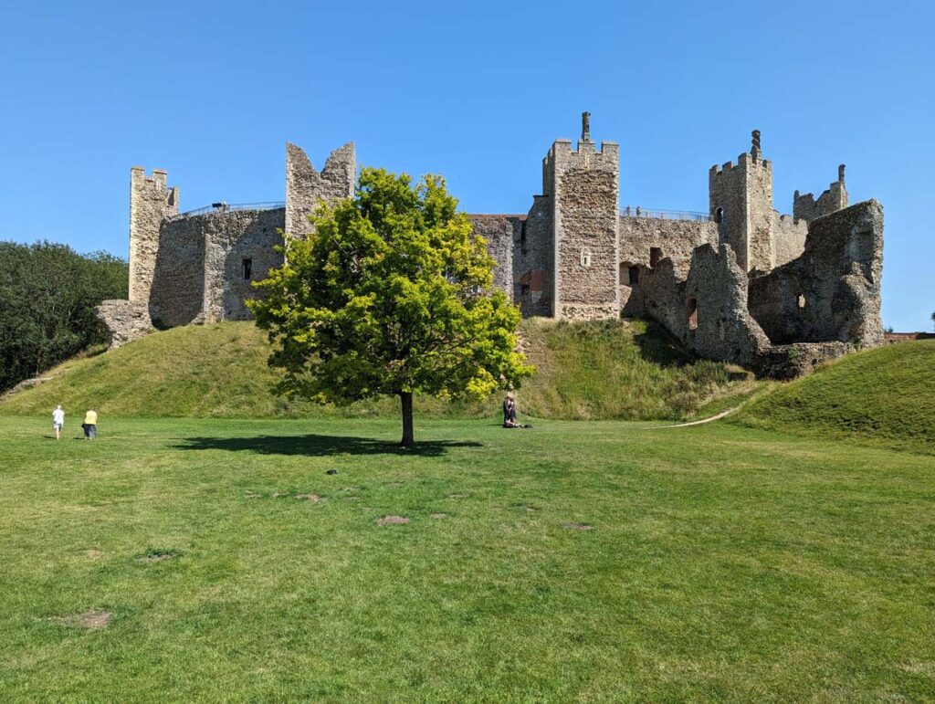 Framlingham Castle from the lower court. A medieval castle with towers is on top of a small grassy hill, with a tree in front. 