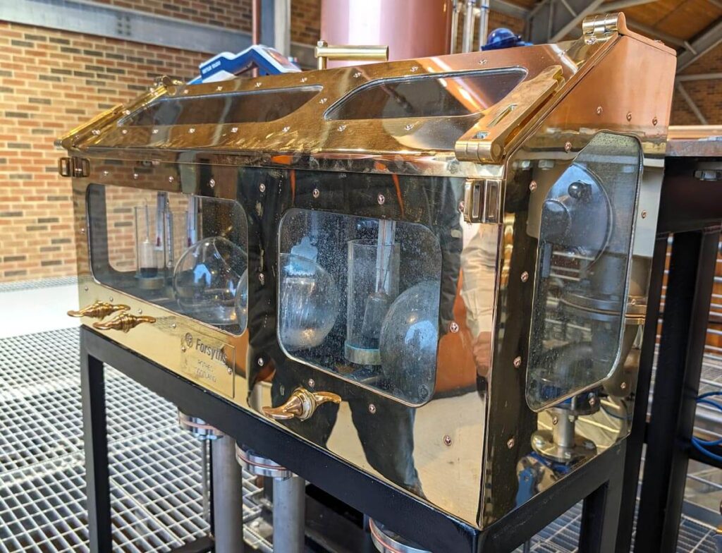A brass cabinet with glass windows and glass flasks inside, used to check the alcohol strength of the spirit coming out of the pot stills.