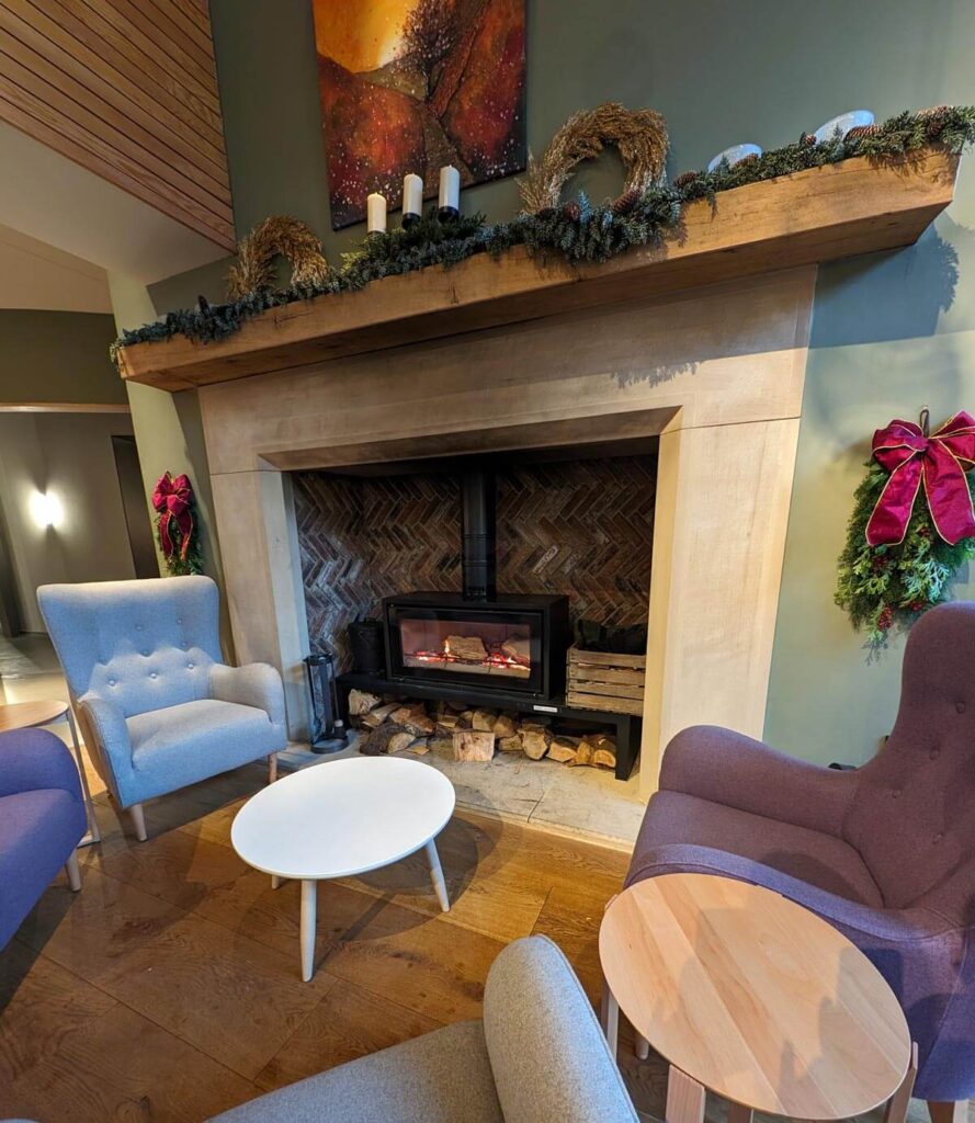 The cosy fire in the bistro. A wood burner is blazing away, with logs stacked up in an inglenook fireplace. Some comfortable chairs are set around the fireplace. 