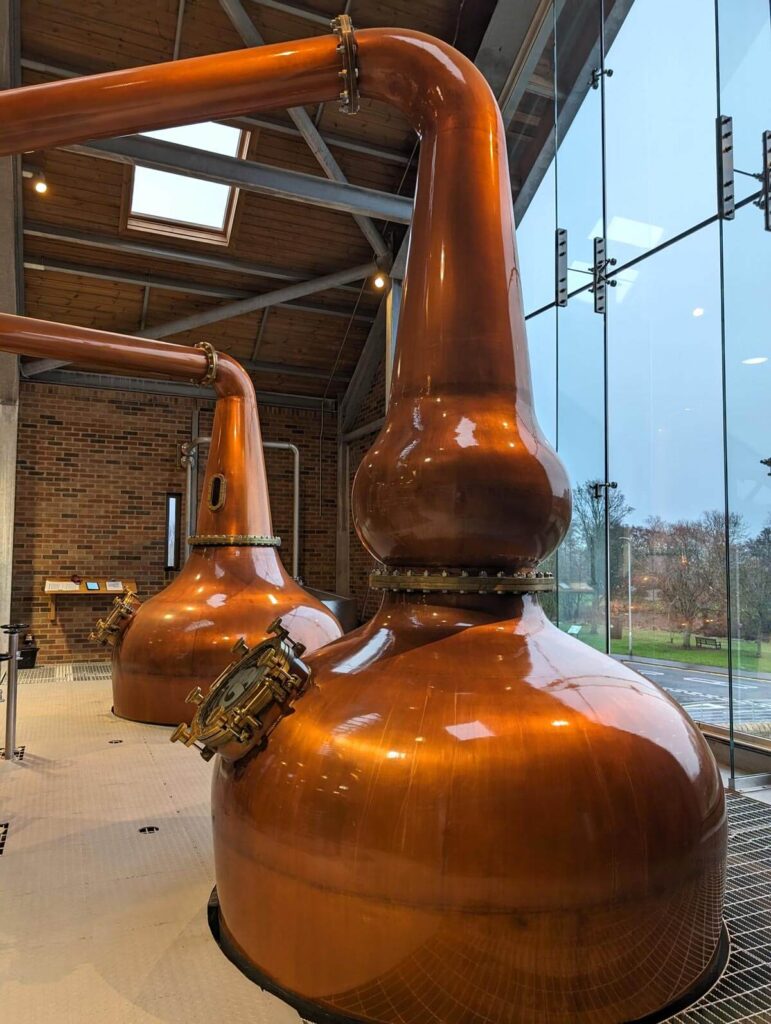 Two large copper pot stills at Ad Gefrin distillery. The one closest to the camera has a bump in the neck; both are twice as tall as a man.