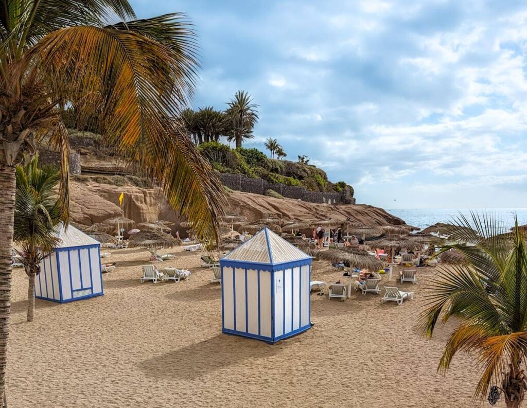 Pretty Playa del Duque in Costa Adeje, Tenerife. Two blue and white striped beach huts sit on a sandy beach, with palm trees and a rocky headland