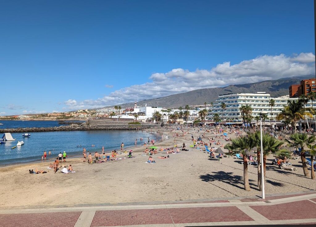 Playa de la Pinta beach in Costa Adeje. The sandy beach curves round and is backed by palm trees and hotels. There are hills and mountains in the background.