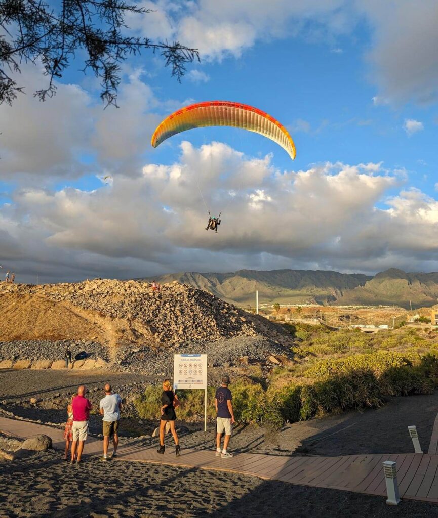 A tandem paraglide with a brightly coloured parachute coming in to land on a beach in Tenerife. The hills where they took off are in the background.