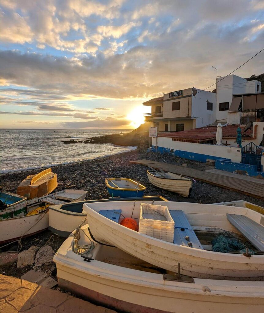 Sunset in La Caleta, a fishing village near Costa Adeje. The sun is setting behind village houses. There are some small white fishing boats on the rocky beach.