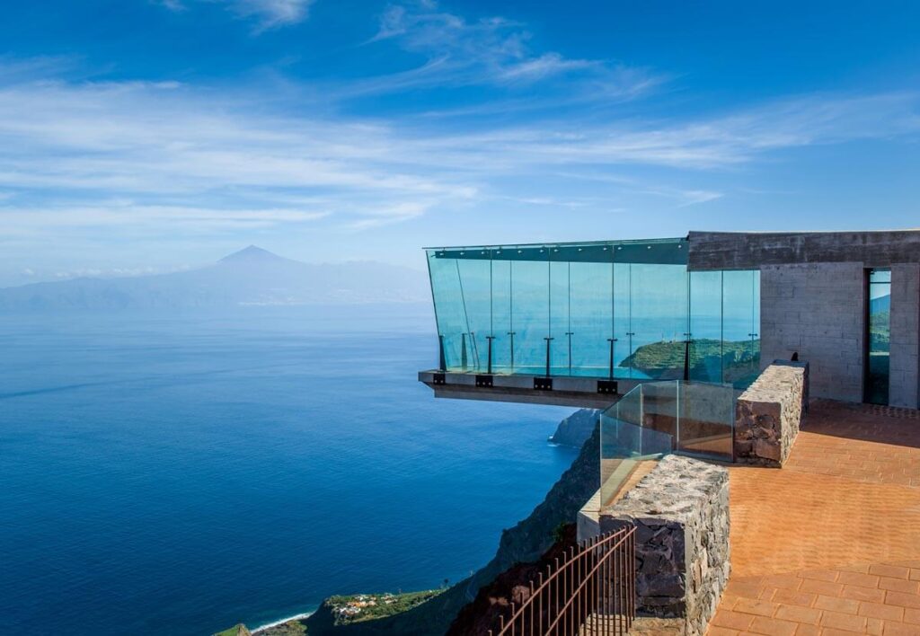 The Abrante glass viewing platform in La Gomera, an island just across the sea from Tenerife. A glass box extends over the edge of the mountain with the sea beyond. The peak of Mount Teide is visible in the distance.
