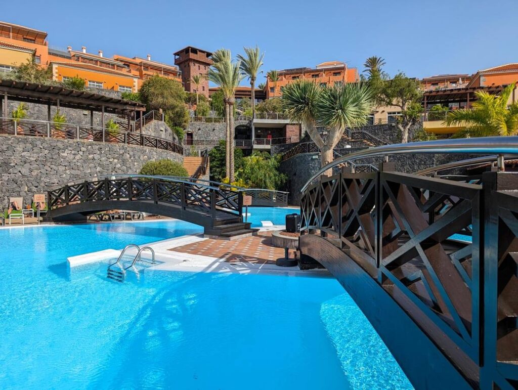 The pool at the Meliá Jardines del Teide hotel. A bright blue pool is crossed by two little bridges. The orange and yellow buildings of the hotel extend up the hill behind.