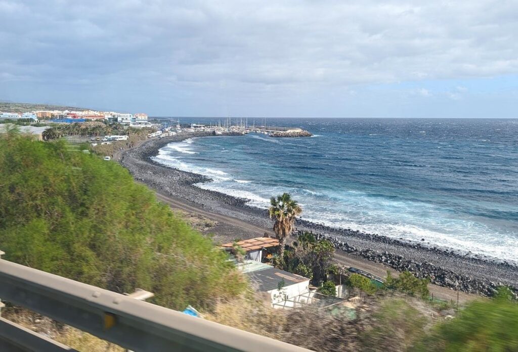 A view from the TF-1 highway from Costa Adeje to the north of the island. The road is close to the ocean; there is a little fishing village with boats in its harbour in the background.