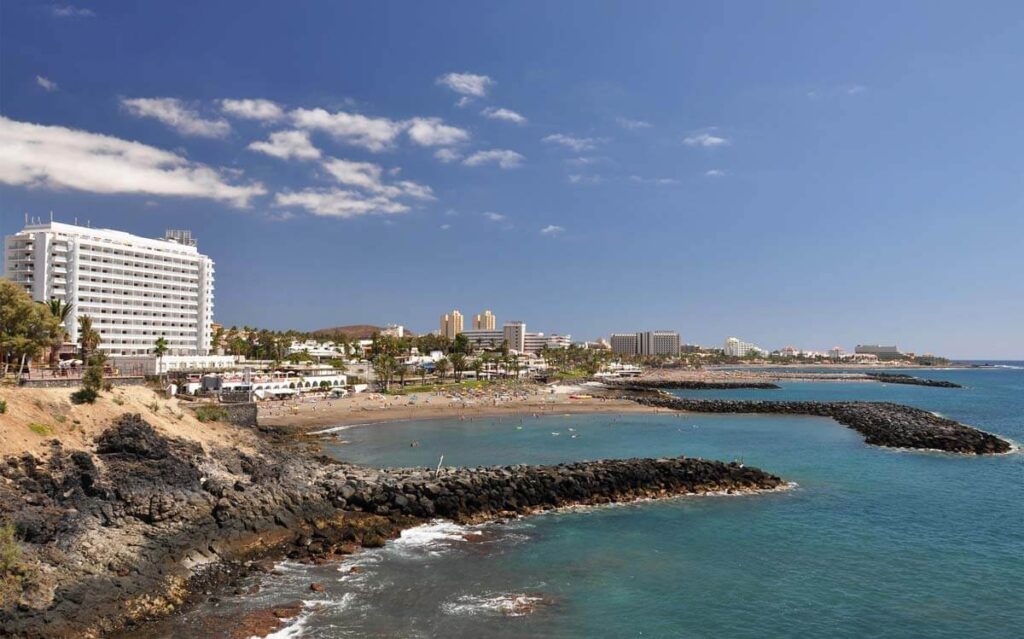 A small beach in Costa Adeje, protected by two large sea walls. There is a large white building - the Playa de las Americas casino - behind the beach.