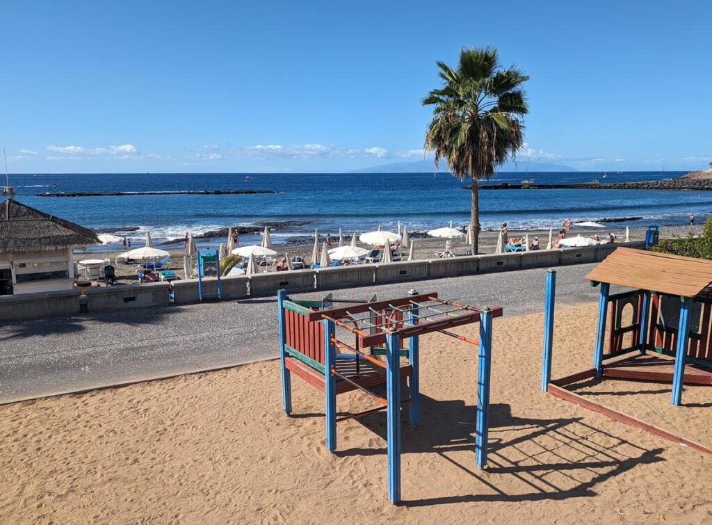 A children's play area on Fañabé beach in Costa Adeje. A climbing frame is in front of a beach promenade, with the beach and the sea beyond.