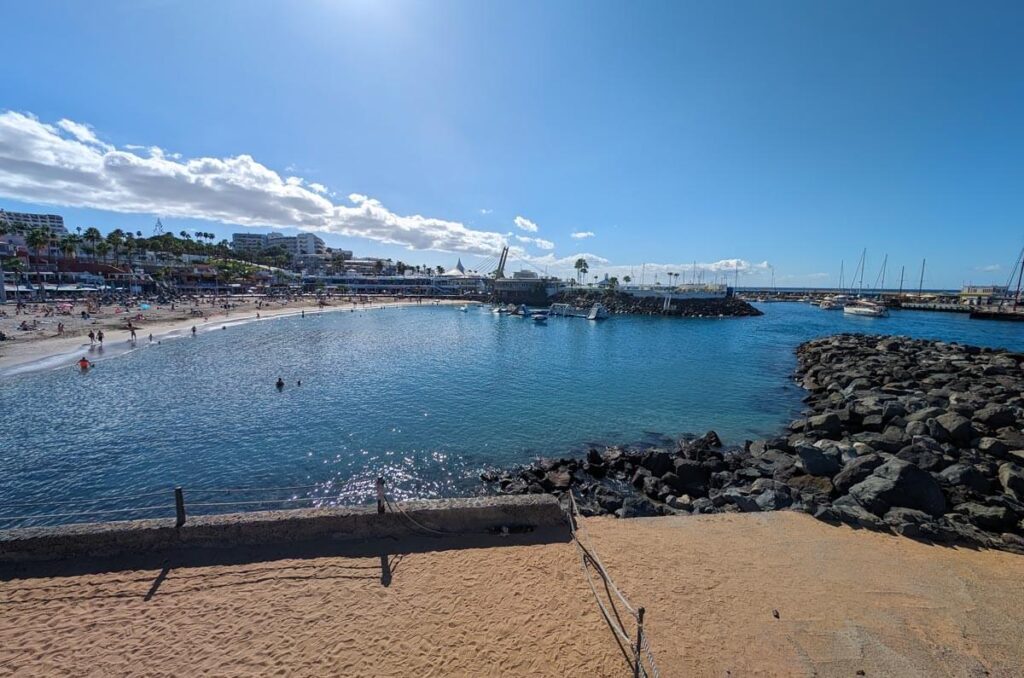 The Pinta beach in Costa Adeje. The beach curves around towards a small harbour. There are boats going in and out of the harbour.