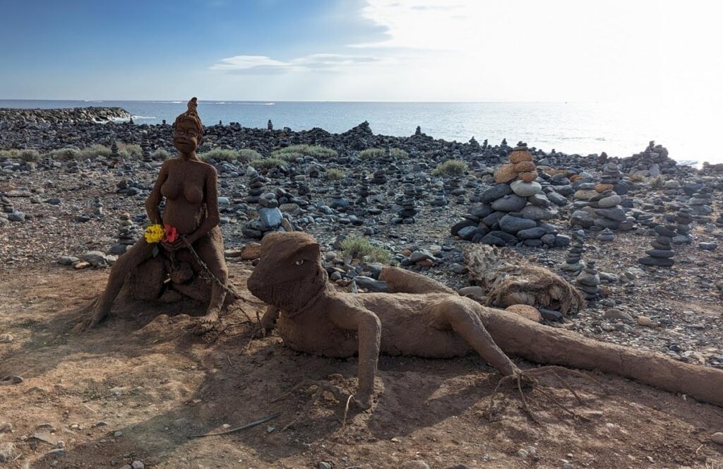 A sand sculpture of a girl and a lizard, with more rock towers and the sea in the background.