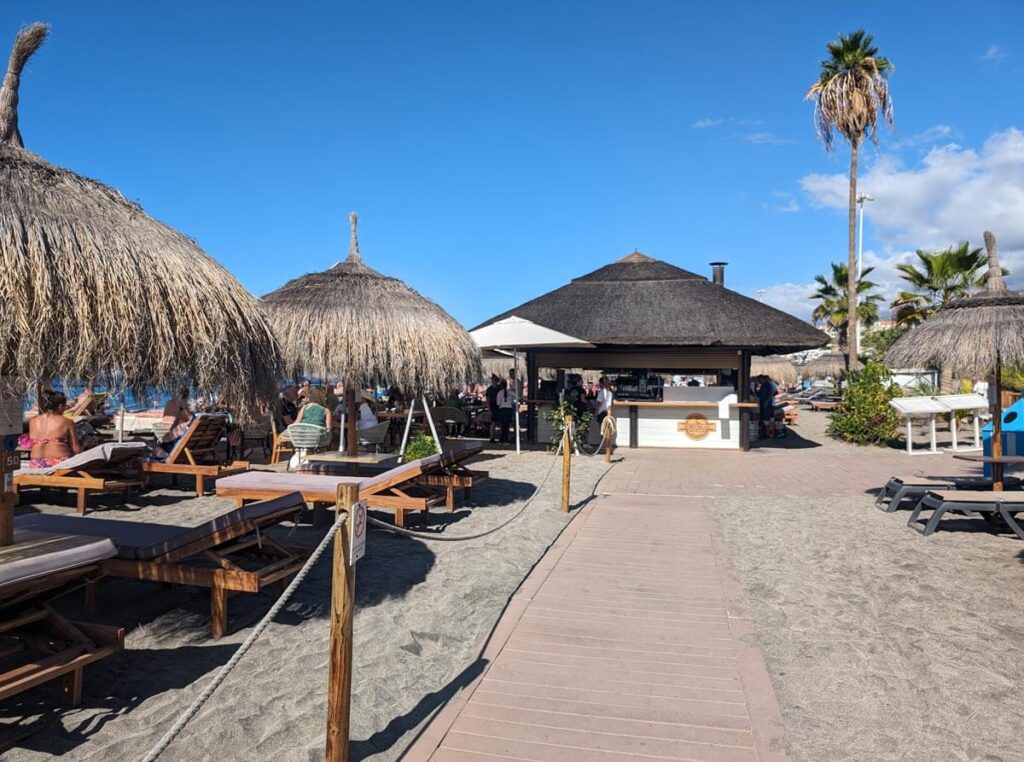 Torviscas Beach Club. A wooden boardwalk leads to a bar with sun loungers and umbrellas