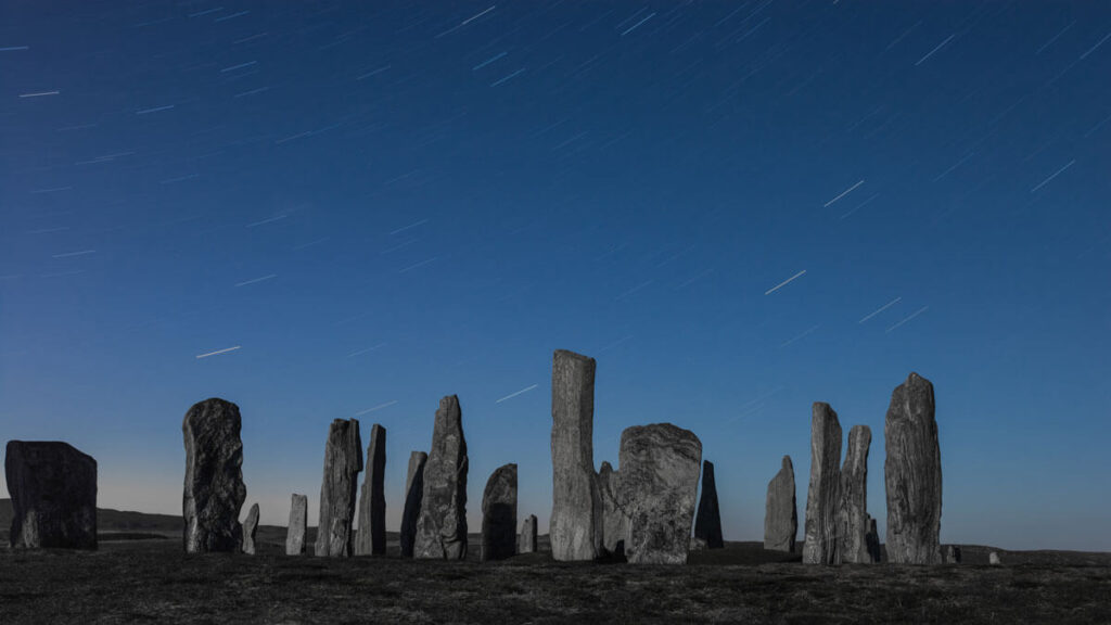 A dark night sky filled with star trails behind irregularly-shaped standing stones