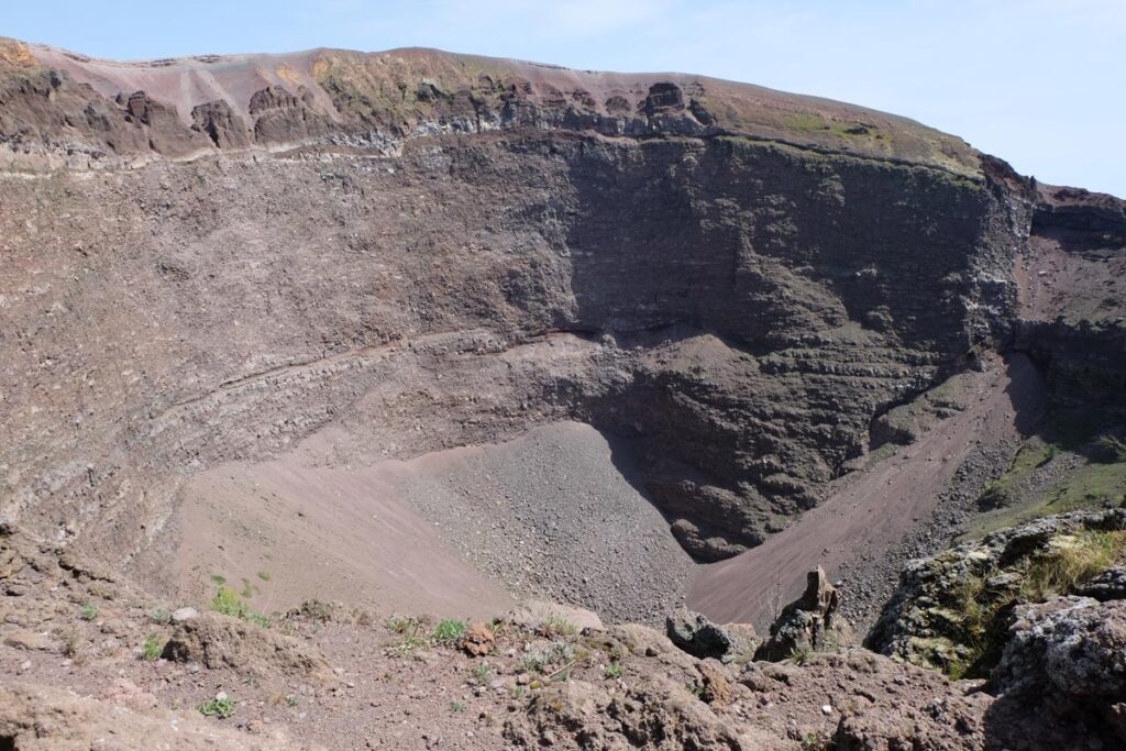 Looking down into the crater of Mount Vesuvius. A giant hole in the ground with red, dusty rocks. It's a mostly barren landscape but there are a few bushes in the crater. 