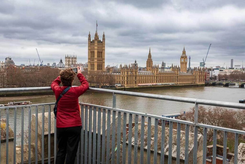 The view from the Garden Museum's tower. A woman is stood at the top of a church tower, holding a camera, with a view of the Houses of Parliament and River Thames.