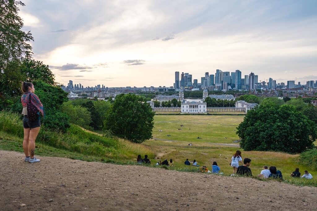 The view from Greenwich park. A woman is stood at the top of a small hill looking at the view, with the white Georgian buildings of the National Maritime Museum in the background and the skyscrapers of Canary Wharf beyond.