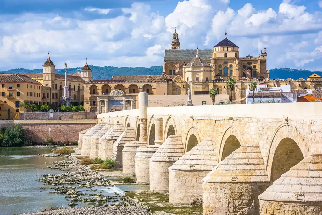 The city of Córdoba. A robust stone bridge with multiple arches crosses a shallow river. There are lots of magnificent old buildings on the other bank, including a huge cathedral.
