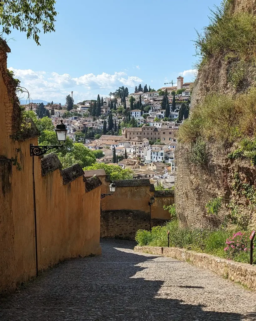 Looking down an old paved street, with a fortification on one side. In the background are the whitewashed houses of the Albaicín area of Granada.