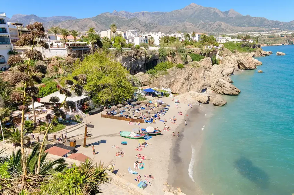 A small beach with sun loungers and a blue sea. Beyond the beach there are rocks. 