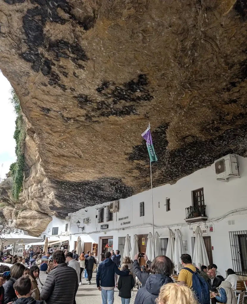 A street with white houses built into and under a huge rocky cliff