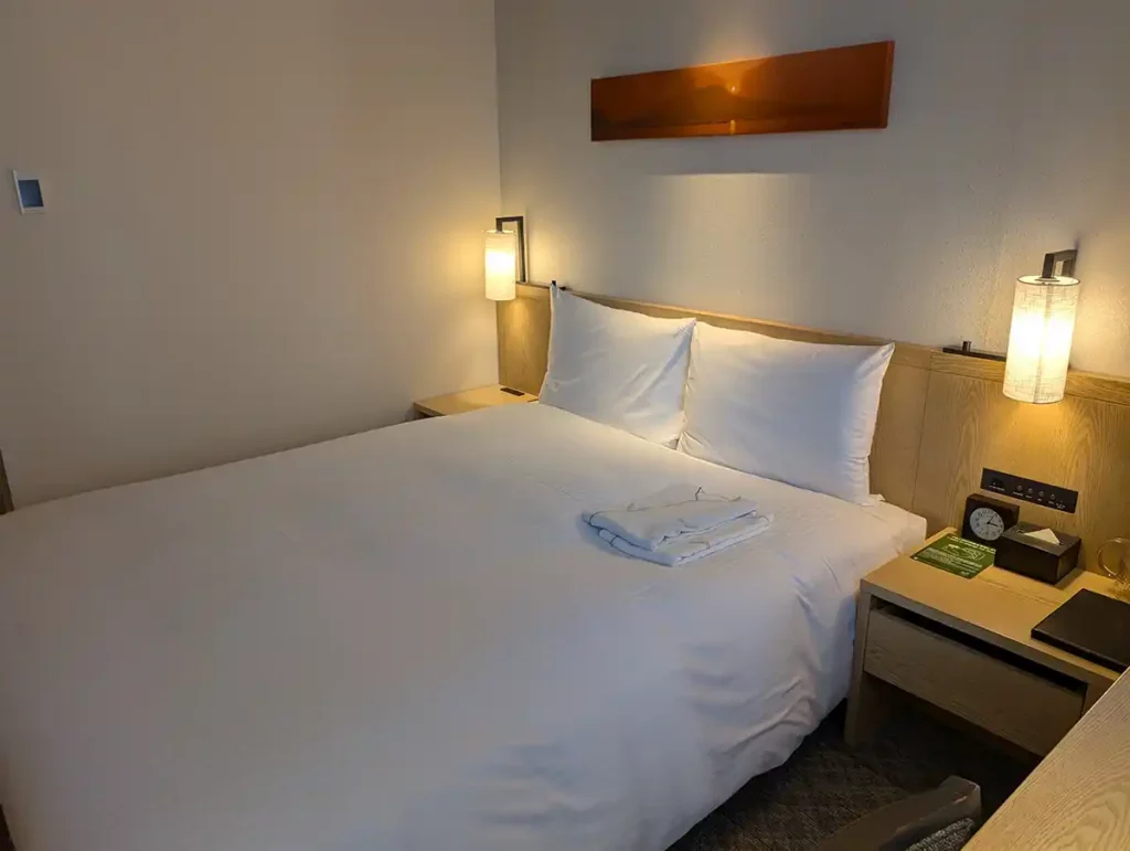 A queen sized bed with white sheets, a lamp on either side and robes folded on the bed at the JR Kyushu Blossom Shinjuku hotel
