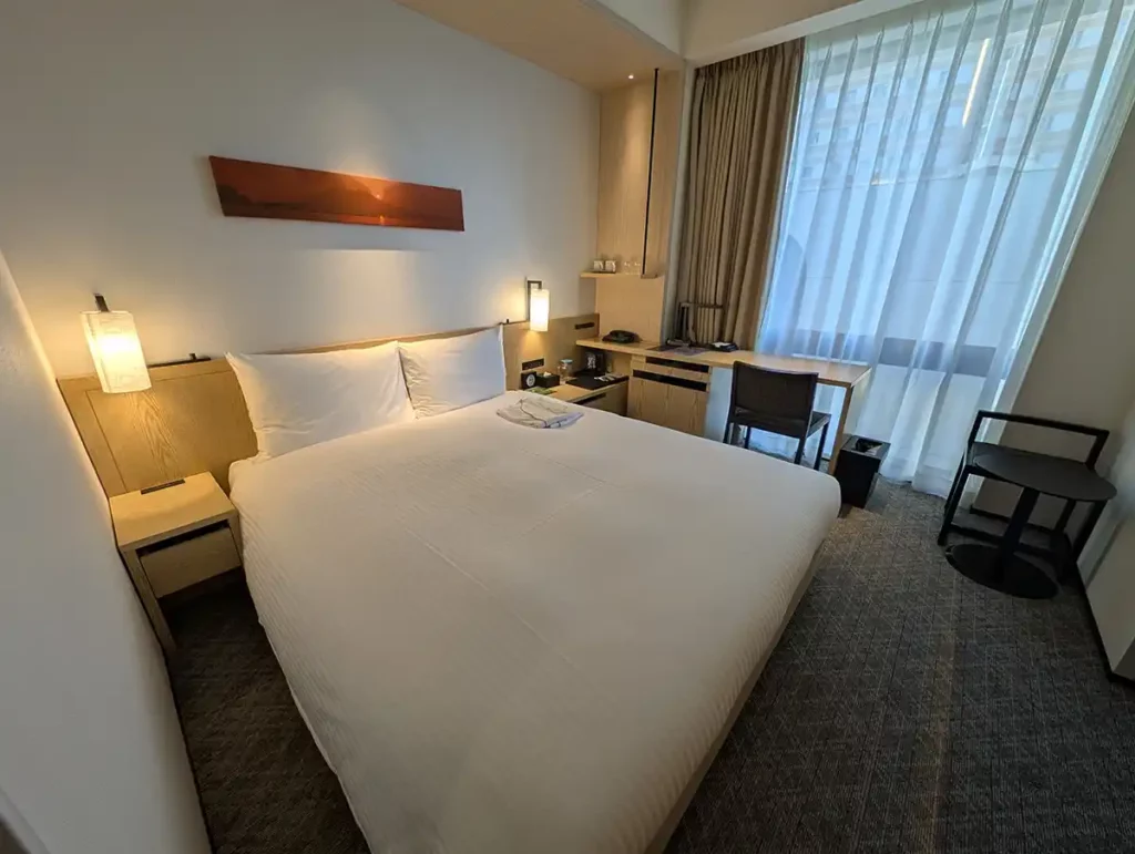 A double queen room at the JR Kyushu Blossom Shinjuku hotel. The bed is queen sized with plenty of space, there's a bedside table and lamp on both sides of the bed and a desk in front of the large, floor to ceiling window.