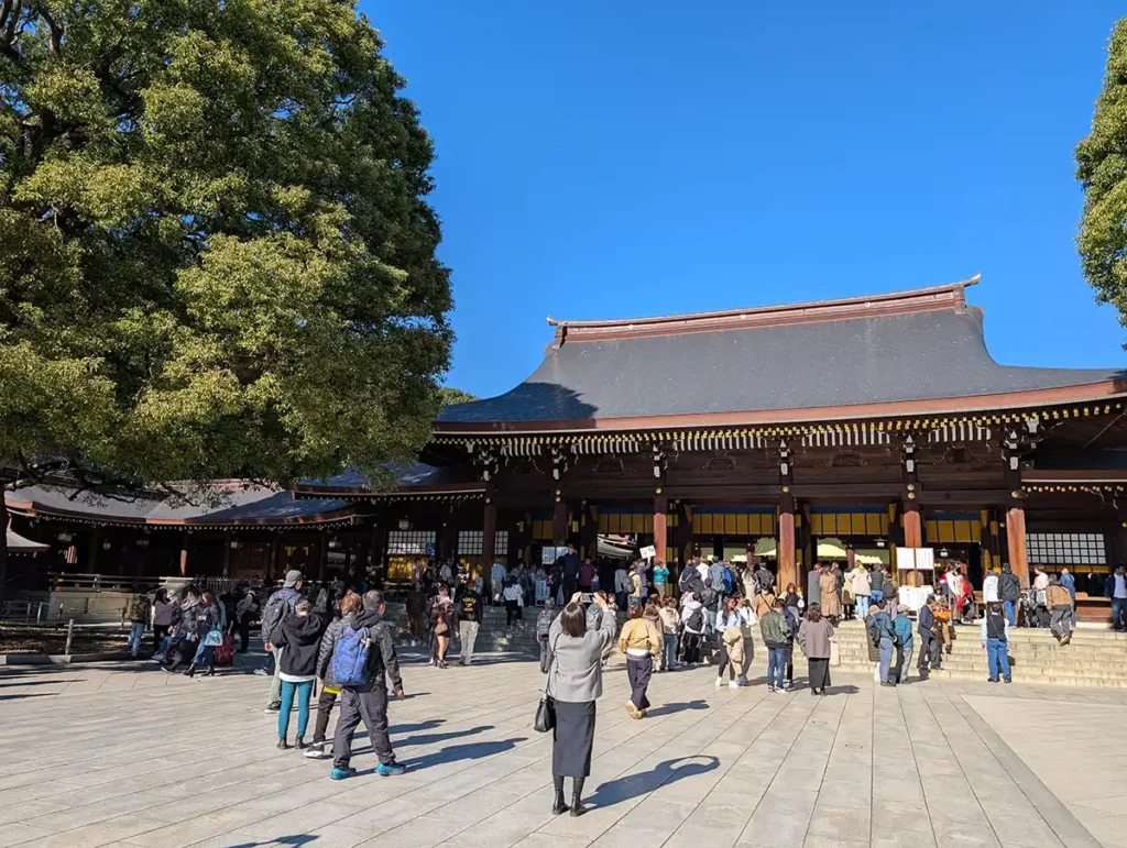 Meiji Jingu Shinto temple. A large courtyard and low temple building with a large tree. People are taking photos.