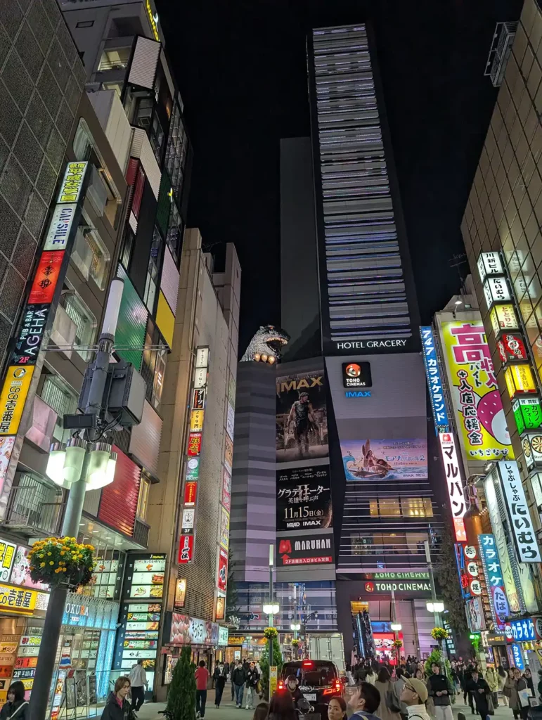 A street in Tokyo with tall buildings and bright lights. There's a Godzilla head on top of one of the buildings.