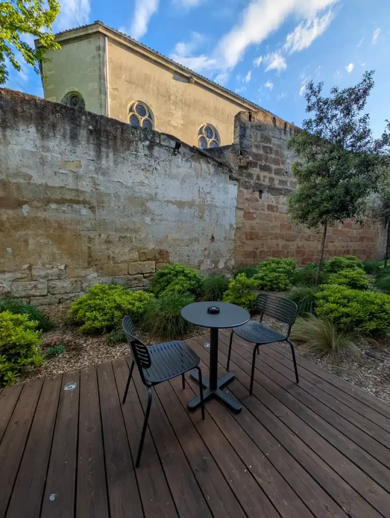A table and chairs on some decking, with an old stone wall and plants in the background