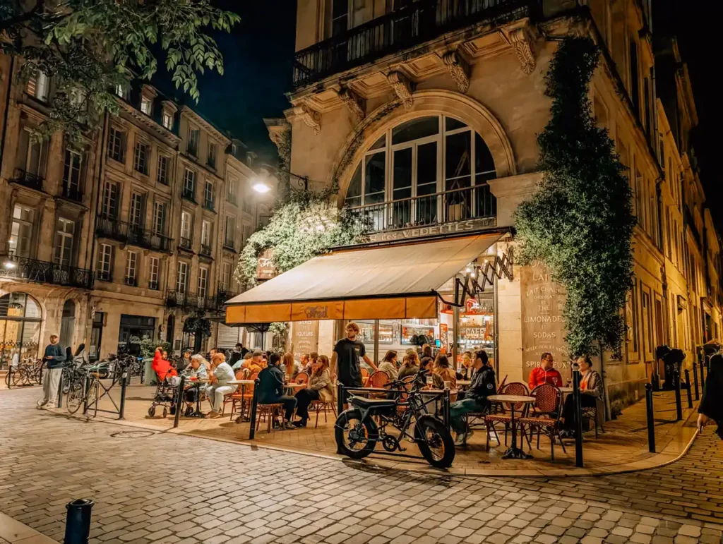 An evening scene of a bar in an old building on a historic street. There are people sat at tables outside.