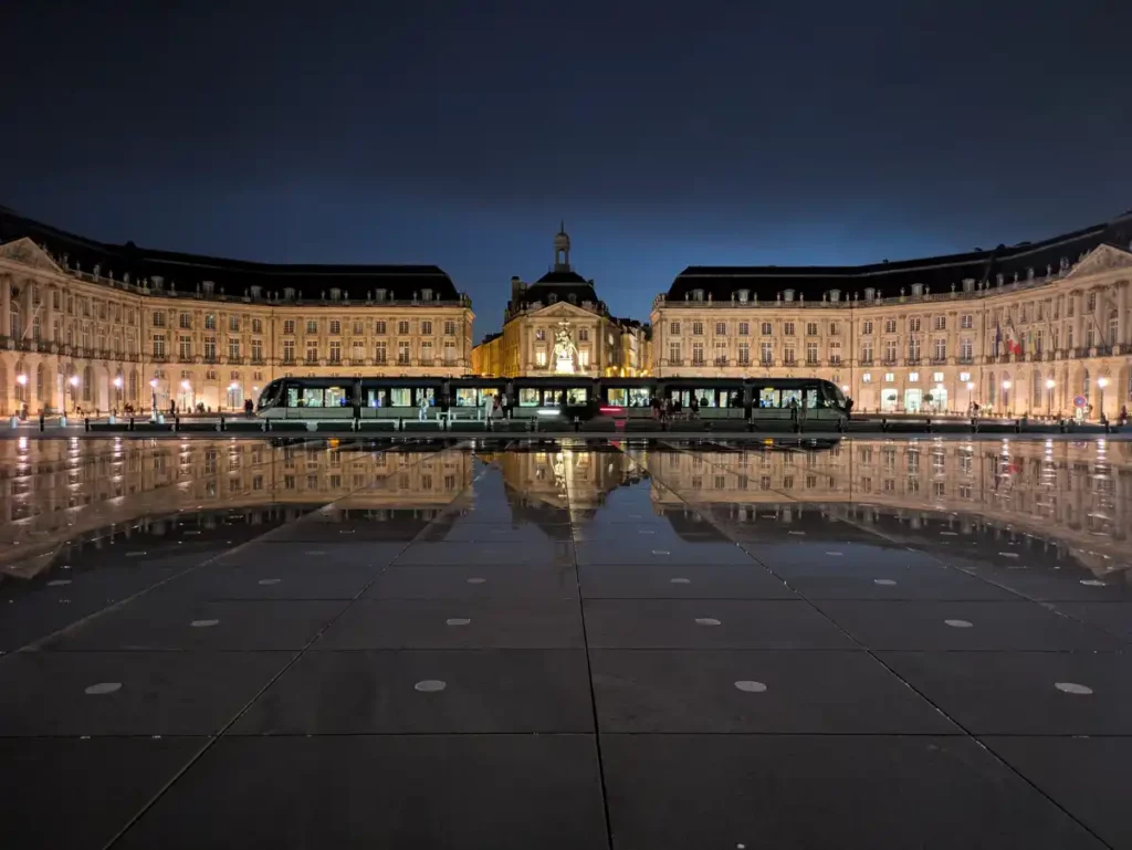 A grand, curving row of buildings at night with a brightly lit tram in front. Both are reflected in the Miroir d'eau, a reflective water feature in Bordeaux.