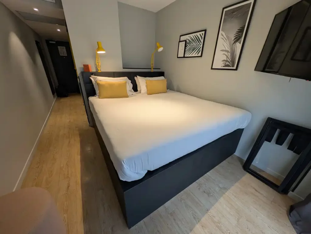 A hotel room with the bed pushed up against one wall. There's a headboard with cubby holes and yellow Anglepoise lamps behind the bed.