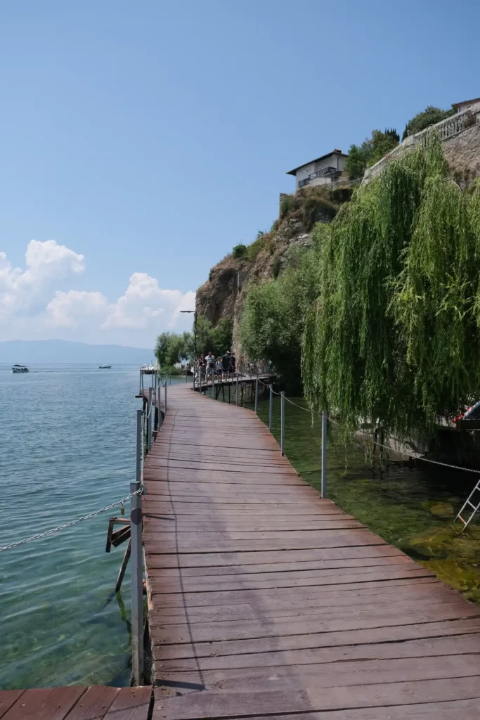 A romantic wooden boardwalk over a blue lake, with cliffs and a weeping willow tree