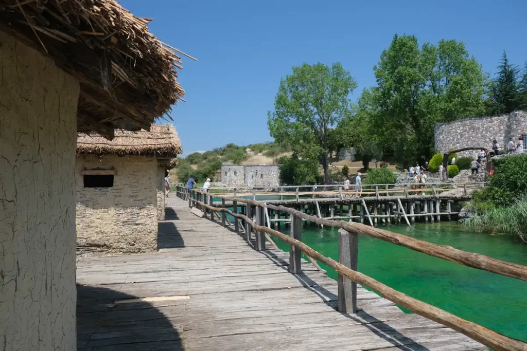 Wooden and straw houses on a wooden platform in the lake. It's connected to the shore by a wooden bridge.